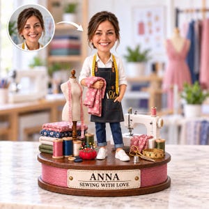 Peut inclure: Une figurine bobblehead personnalisée représentant une couturière portant un tablier noir sur une chemise blanche et un jean bleu, tenant un tissu floral rose. La base en bois porte une plaque ANNA SEWING WITH LOVE et des accessoires de couture.