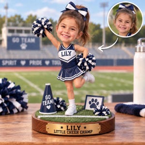 Pode incluir: Uma figura bobblehead personalizada de uma jovem líder de torcida vestindo um uniforme azul-marinho e branco com o nome LILY no peito. A figura segura pompons azul-marinho e brancos, sobre uma base de grama com um megafone e uma placa.