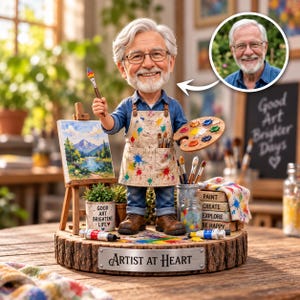 May include: A custom bobblehead figurine of an older person with white hair and glasses, wearing a paint-splattered apron over a blue shirt. The figure holds a paintbrush and palette, standing on a wood base labelled ARTIST AT HEART with art supplies.