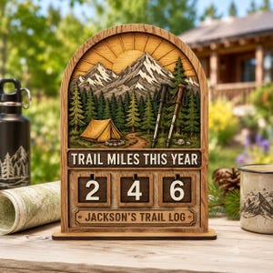 May include: A wooden arched sign featuring a mountain and forest scene with a yellow tent and two hiking poles. The sign displays the text "TRAIL MILES THIS YEAR" above three rotating number blocks showing "246" and "JACKSON'S TRAIL LOG".