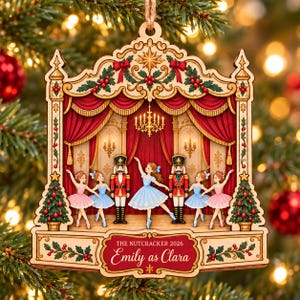 May include: A wooden Christmas ornament shaped like a theatre stage featuring a Nutcracker ballet scene with dancers and nutcracker soldiers. The text reads THE NUTCRACKER 2026 Emily as Clara. The design includes red curtains, a chandelier, and holly.