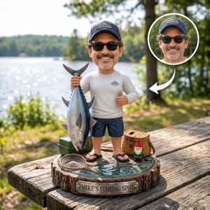 May include: A custom bobblehead figure of a smiling person wearing a blue cap, sunglasses, and a long-sleeve shirt, holding a large silver fish. The base features a small garden gnome, a fishing net, a green tackle box, and a plaque reading MIKE'S FISHING SPOT.