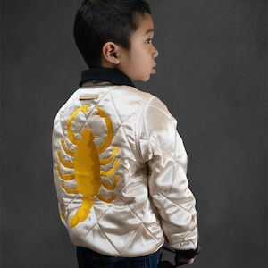 May include: Cream-colored quilted jacket with a large yellow scorpion embroidered on the back. The jacket has a black collar and cuffs. The child is wearing blue jeans. The jacket is a stylish outerwear piece.