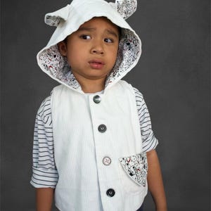 May include: A white corduroy vest with four black buttons and a pocket with a black and white pattern. The vest is worn over a striped shirt. A matching white hood with ears and a patterned lining completes the outfit.
