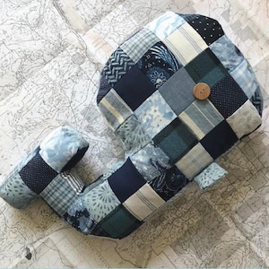Puede incluir: Un juguete de ballena de patchwork en tonos azules, marino y blanco, con un botón como ojo. El juguete está hecho de varios cuadrados de tela, creando un patrón similar a un edredón. El texto "PATCHWORK WHALE TOY" y "A quilt pattern by Kirious Design Co." son visibles.