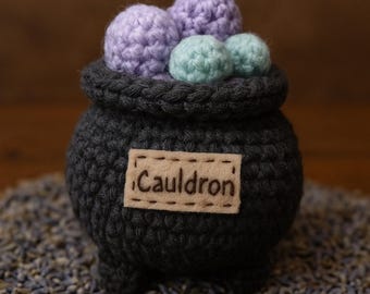 Op de afbeelding: Een gehaakte ketel met een label met de tekst "Cauldron" en gevuld met paarse en groene garenballen.