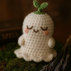 Puede incluir: Un fantasma blanco de crochet con un brote verde en la cabeza. El fantasma tiene una cara sonriente con los ojos cerrados y mejillas rosadas. Está sentado en un lecho de musgo verde con ramitas de lavanda en primer plano.