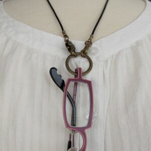 May include: A black cord necklace with a gold-toned clasp and a pair of reading glasses attached to it. The glasses have a burgundy frame and clear lenses.