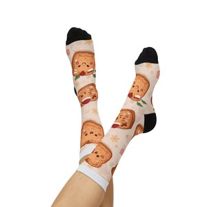 Könnte beinhalten: Ein Paar Socken mit einem sich wiederholenden Muster von lächelnden Toast-Charakteren mit roten und grünen Akzenten. Die Socken haben schwarze Fersen und Zehen sowie einen weißen Bund. Der Hintergrund ist hellbeige mit kleinen Sternformen.