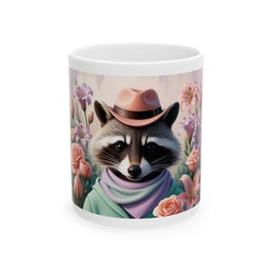 Könnte beinhalten: Eine weiße Keramiktasse mit einer bunten Illustration eines Waschbären, der einen rosafarbenen Cowboyhut und einen lila Schal trägt. Der Waschbär ist von rosa und lila Blumen umgeben.