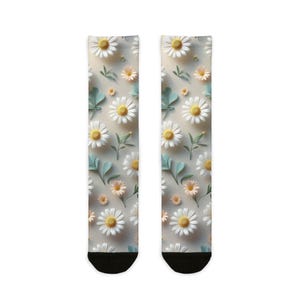Könnte beinhalten: Socken mit einem sich wiederholenden Muster aus weißen Gänseblümchen mit gelben Zentren, kleinen pfirsichfarbenen Blüten und hellblauen Blättern. Die Socken haben schwarze Zehen und Fersen, vor einem neutralen Hintergrund.