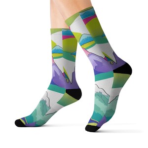 May include: A pair of socks featuring a colorful mountain landscape design. The socks display a geometric pattern with shades of purple, green, blue, and pink. The socks have black heels and toes, and are worn on a person's feet.