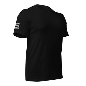May include: Black crew neck t-shirt with a white American flag design on the left sleeve. The word "FREEDOM" is printed below the flag. The shirt is made of a soft material.