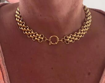 Puede incluir: Un collar dorado con un diseño de cadena de tres hilos. El collar presenta una serie de eslabones interconectados, creando una apariencia audaz y texturizada. El cierre es grande y circular. Una pieza de joyería llamativa.