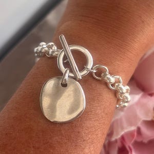 Può includere: Un braccialetto d'argento con chiusura a toggle e un ciondolo rotondo. Il braccialetto presenta una catena di piccole perline rotonde e viene indossato su un polso. Lo sfondo include un fiore rosa.