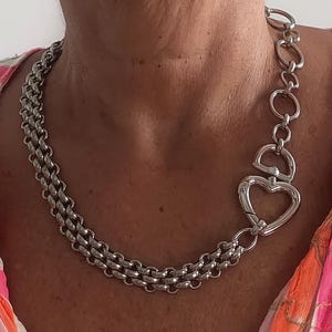 Può includere: Una collana argentata con un design a catena a doppia fila. La collana ha una chiusura a forma di cuore e una serie di maglie di collegamento sul lato. Lo sfondo mostra un top con motivi rosa, arancioni e bianchi.