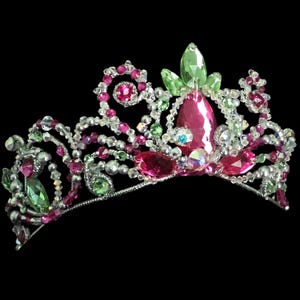 May include: A silver-toned tiara featuring a decorative arrangement of faceted pink and light green crystals. The metal frame is intricately wired with small clear beads and larger teardrop-shaped stones, creating a symmetrical, ornate headpiece.