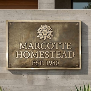 May include: A rectangular bronze plaque with a polished gold-toned border and four corner screws. The center features a raised floral emblem above the text MARCOTTE HOMESTEAD and EST. 1980 in a classic serif font, set against a dark, textured background.