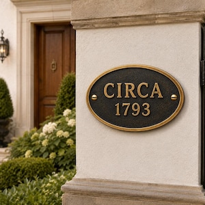 Puede incluir: Una placa de forma ovalada con un fondo oscuro texturizado y un borde dorado en relieve. El texto "CIRCA 1793" está grabado en letras doradas en el centro. La placa está fijada a un pilar de piedra de color claro con dos tornillos visibles.