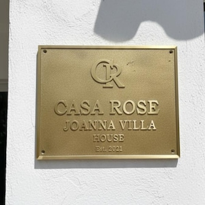 Pode incluir: Placa retangular dourada com as palavras "CASA ROSE JOANNA VILLA HOUSE Est. 2021" em uma fonte serifada. A placa também apresenta as letras entrelaçadas "CR" no topo. A placa está montada em uma parede branca texturizada.
