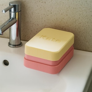 May include: A stacked soap dish with a yellow lid and a pink base, with the name "Kate" embossed on the lid. The dish has a ribbed design on the sides. The soap dish is on a white sink next to a chrome faucet.