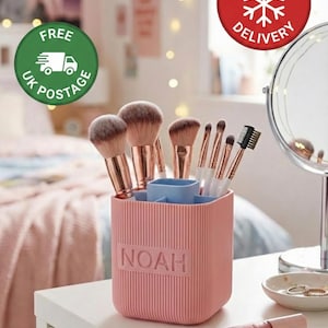 Puede incluir: Un soporte rosa para brochas de maquillaje con el nombre "NOAH" en relieve en la parte delantera, lleno de varias brochas de maquillaje. El soporte está sobre una superficie blanca, con un espejo y otros productos de belleza en el fondo. La imagen también incluye una pegatina de "Custom Colour Options".