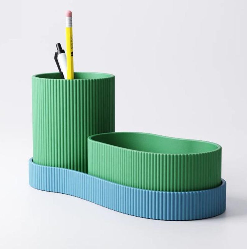 May include: A green and blue desk organizer with a ribbed texture. The organizer has two sections: a tall, cylindrical cup and a smaller, oval-shaped container. A yellow pencil and black pen are inside the tall cup. The base is blue.