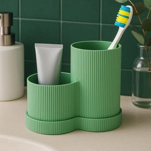 May include: A green toothbrush holder with a ribbed design, holding a white toothbrush with blue and yellow bristles. A tube of toothpaste sits in a smaller compartment. The holder is on a white surface.