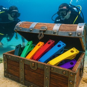 Könnte beinhalten: Eine offene hölzerne Schatzkiste unter Wasser, gefüllt mit bunten, geometrischen Objekten. Taucher in schwarzen Neoprenanzügen und Tauchausrüstung sind im Hintergrund. Die Truhe steht auf sandigem Meeresboden.
