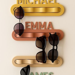 May include: Four sunglasses holders in yellow, terracotta, beige, and green, each with a name: MICHAEL, EMMA, JAMES. Each holder has a curved slot to hold sunglasses. Several pairs of sunglasses are displayed. The holders are a stylish storage solution.