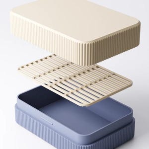 May include: An exploded view of a rectangular lunch box. The lid and base are a light beige color with vertical ribbed detailing. The interior container is a muted blue, and a beige slotted tray sits inside.