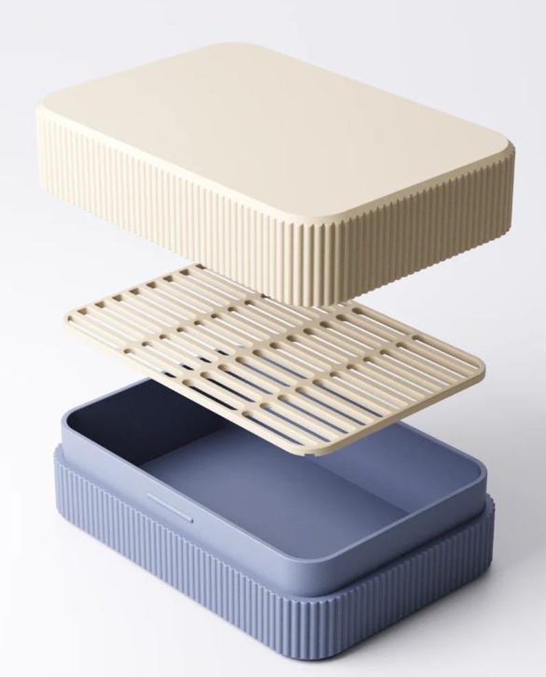 May include: An exploded view of a rectangular lunch box. The lid and base are a light beige color with vertical ribbed detailing. The interior container is a muted blue, and a beige slotted tray sits inside.