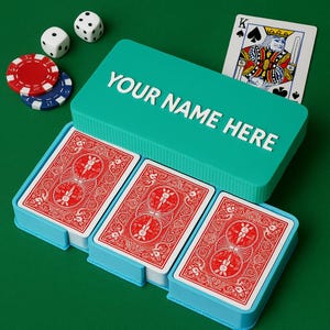 May include: A teal playing card case with the text "YOUR NAME HERE" in white lettering. The case holds three decks of cards with red backs, a King of Spades card, two dice, and poker chips. The background is a solid green.
