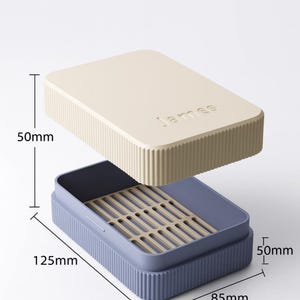 May include: A rectangular storage box with a cream-colored lid and a blue base. The lid has the name "James" embossed on it. The box is 50mm high, 125mm long, and 85mm wide. The interior has a beige tray with slots.