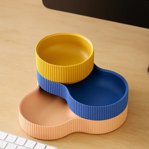 May include: A three-section pet bowl in yellow, blue, and peach. Each section is a circular bowl with vertical ridged sides. The bowls are stacked in a cloverleaf shape. The product is on a wooden desk with a keyboard in the foreground.