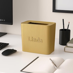 Puede incluir: Un organizador de escritorio rectangular dorado con detalles acanalados verticales. El nombre "Linda" está grabado en relieve en la parte delantera. El organizador está sobre un escritorio blanco, junto a un ratón de ordenador negro y un cuaderno.