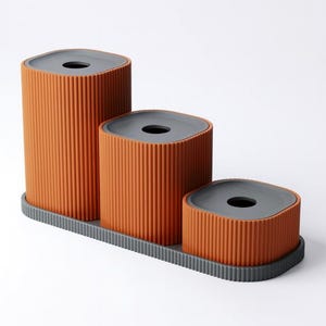 May include: Three orange cylindrical containers with gray lids and a ribbed texture, arranged in a stepped configuration on a gray rectangular base. The containers have a hole in the center of each lid.