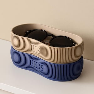 Può includere: Un paio di occhiali da sole in un contenitore beige con la scritta "HIS", impilato su un contenitore blu con la scritta "HERS". Entrambi i contenitori presentano dettagli a coste verticali. Gli occhiali da sole hanno montatura e lenti scure.