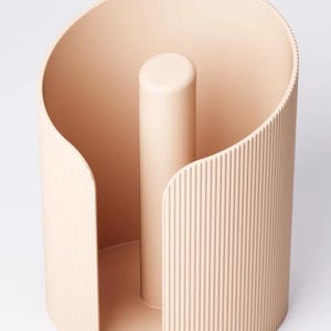 May include: A beige paper towel holder with a cylindrical design. It features a central post and a curved, ribbed outer surface. The holder has a minimalist, modern aesthetic with a smooth, matte finish. Designed to hold a standard roll of paper towels.