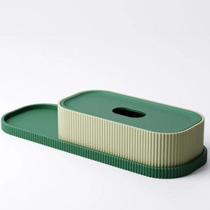 May include: A green and beige tissue box with a matching tray. The tissue box has a ribbed texture and a dark green lid with an oval opening. The tray is a darker shade of green and has a ridged edge.