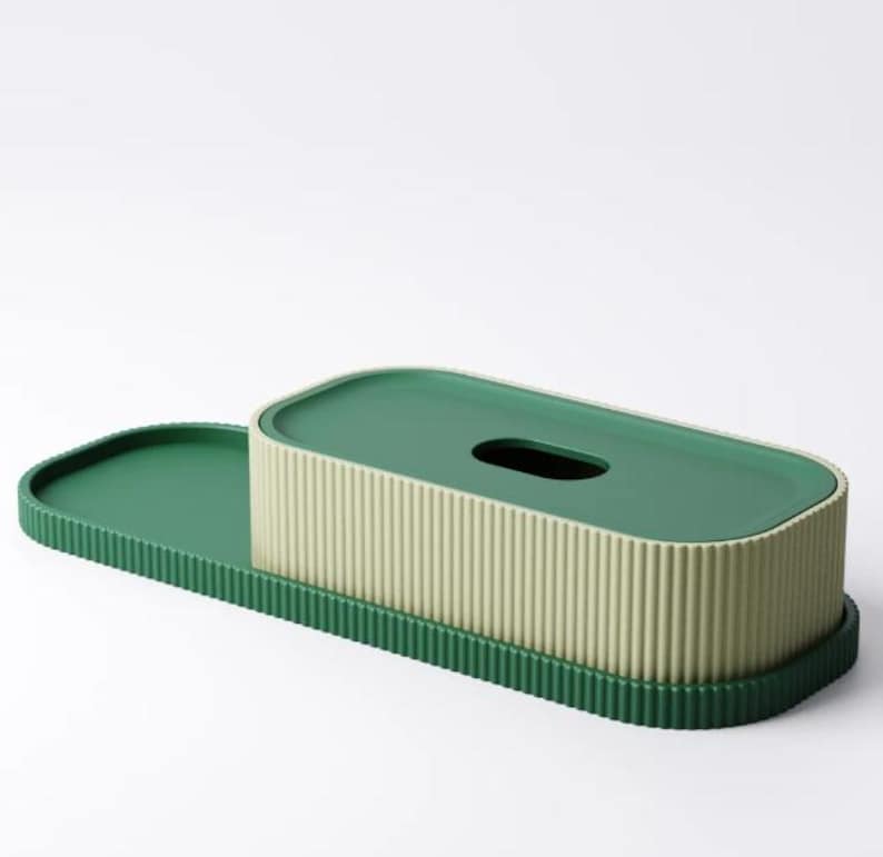 May include: A green and beige tissue box with a matching tray. The tissue box has a ribbed texture and a dark green lid with an oval opening. The tray is a darker shade of green and has a ridged edge.