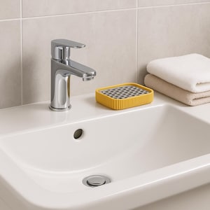 May include: A contemporary bathroom sink featuring a chrome tap and a yellow soap dish. The soap dish has a grey grid pattern and sits on the white countertop. Two folded towels are stacked on the right.