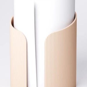May include: A cylindrical paper towel holder with a light peach-coloured, ribbed exterior. The holder has a curved design that partially wraps around a roll of white paper towels, creating a modern and minimalist aesthetic.