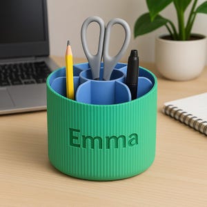 Puede incluir: Un organizador de escritorio verde y azul con el nombre "Emma" en relieve en la parte delantera. El organizador contiene un lápiz amarillo, unas tijeras grises y un bolígrafo negro. El organizador es cilíndrico con ranuras verticales.