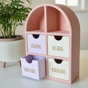 Könnte beinhalten: Ein rosa, bogenförmiges Aufbewahrungselement mit vier Schubladen, die mit "SEWING", "BEADS", "THREADS" und "RIBBONS" in goldfarbenen Buchstaben beschriftet sind. Das Element ist aus Holz gefertigt und hat eine hellrosa Oberfläche. Eine Zimmerpflanze in einem weißen Topf befindet sich im Hintergrund.