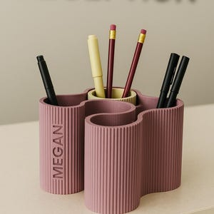 May include: A mauve desk organizer with a wavy design, holding pens and pencils. The organizer has vertical ribbed detailing and the name "MEGAN" is printed on the side. The word "RECEPTION" is blurred in the background.