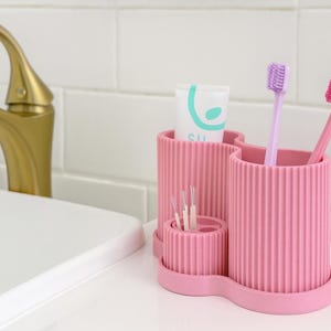 Op de afbeelding: Een roze tandenborstelhouder met een geribbeld ontwerp, met twee tandenborstels en een tube tandpasta. De houder staat op een wit oppervlak, met een gouden kraan op de achtergrond. Er is ook een klein compartiment voor tandenstokers.