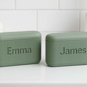 May include: Two sage green rectangular boxes with lids, each embossed with a name: "Emma" and "James". The boxes have a ribbed texture and are on a white surface. A white towel and a white soap dispenser are in the background.