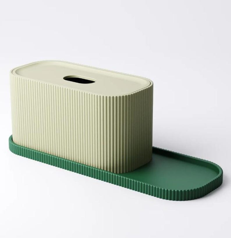May include: A tissue box with a ribbed, light green exterior and a matching lid. The box sits on a dark green tray with a ribbed edge. The design is modern and minimalist.