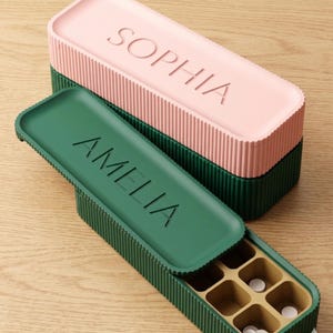 Puede incluir: Organizadores de pastillas rectangulares verdes y rosas con laterales acanalados. La caja superior es rosa con el nombre "SOPHIA" grabado. La caja inferior es verde con el nombre "AMELIA" grabado. La caja abierta contiene pastillas.